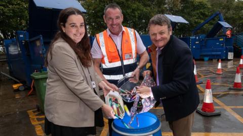 A woman and two men empty plastic containers and a bag full of disposable vape pens into a blue recycling bin at a tip site, with waste containers and traffic cones in the background.