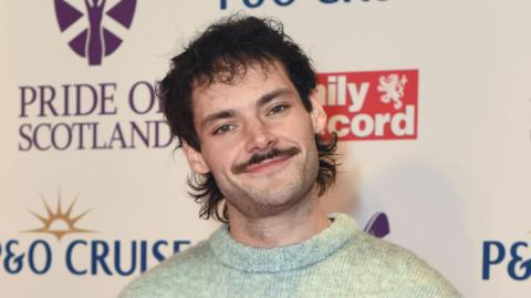 A man with a brown mullet and moustache wearing a light green knitted jumper. He is standing in front of a white backdrop.