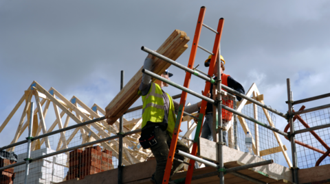 Two men on scaffolding on a house, one holds wood on his shoulder while talking to the other