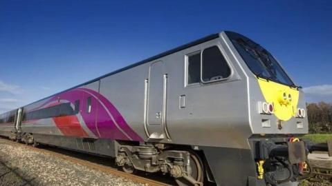 A grey train with swirls of purple and red is stationary on the train line.