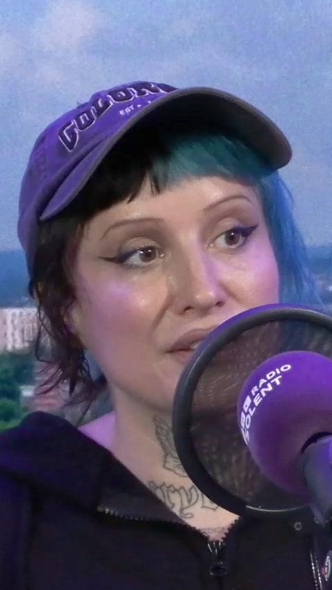 Rox Pink sits in the Radio Solent studio with a blue background. She has her hair tied back in a ponytail with a fringe. Half her hair is died blue and half is black. She has a black peaked cap on her head and tattoos on her neck and wears a black zipped up hoodie.