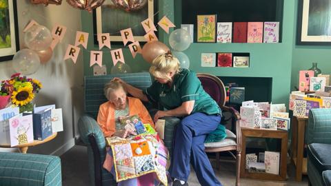 Bertha Preston sitting in a green arm chair in the care home, with a quilted blanket and a card from King Charles III resting on her lap. She is wearing a green top underneath an orange cardigan. Beside her is her carer, Jane Rogers, sitting with her legs crossed, wearing a green shirt and blue trousers. Jane is gently stroking the hair away from Bertha's face. They are surrounded by cards, happy birthday banners, balloons and flowers. 