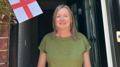 Maxine Simeone, who has shoulder length blonde hair and a green top, smiles at the camera from inside the doorway of a terraced house. A red and white England flag is to her right. 
