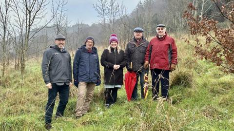 Five people standing beside each other smiling, on a plot of land which has been secured in the new deal. They are surrounded by trees which have no leaves.