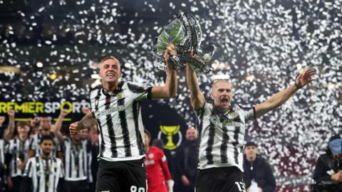 Two footballers in black and white striped kit lift the Scottish League Cup, with confetti in the background