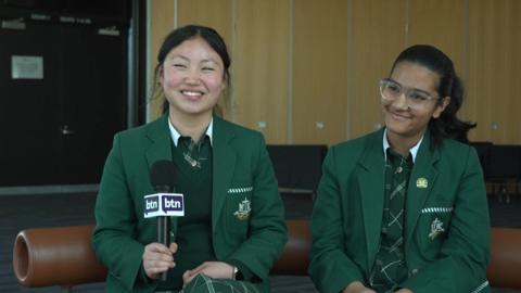 Two teenagers, wearing school uniform, hold a microphone and smile whilst being interviewed