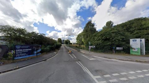 General view of the junction of a road with hedges and trees on both sides. On the left there is a dark blue banner with white writing on it. On the right, near a road sign, there is a white and green banner that also has writing on it