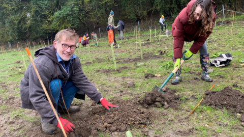 A smiling man wearing red gardening gloves and warm clothes kneels by a pile of soil, ready to plant a young sapling in a muddy grassy field. A woman digs another hole next to him with a spade.