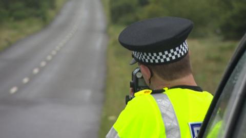 A police officer next to a single carriageway rural road with a speed gun. 
