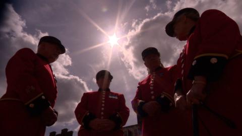 The sun beams from a blue sky with little fluffy clouds onto a group of four retired army soldiers who are wearing scarlet red jackets with shining gold buttons down the front