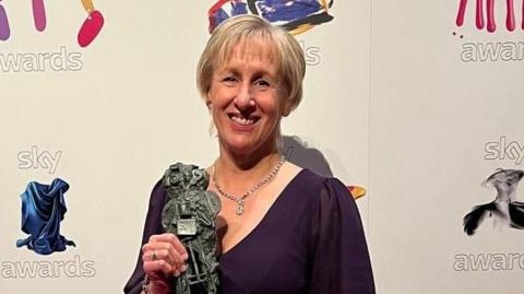 A woman in a purple dress holding an award 

She has light hair and is smiling