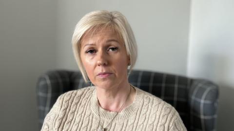 An image of Catherine sitting in an armchair in her living room, looking directly to the camera