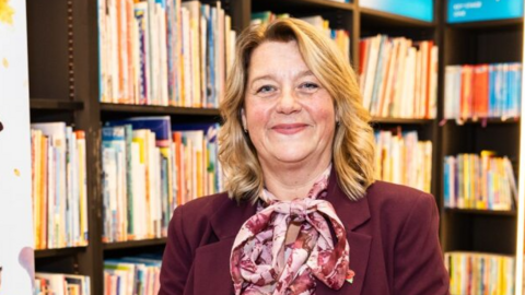 Julie Broadbent is stood in the middle of the image holding an award in the middle with both hands. She has shoulder length dark blonde hair and is wearing a pink blouse and burgundy suit jacket. She is stood in front of a book case.