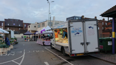 The Wednesday market at Sevenoaks being held in a car park next to the town's bus station. A fish van is in the foreground, with several stalls set up either side of the entrance and exit road.