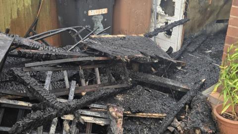 A burnt-down shed reduced to a pile of charred wooden planks on the ground. Next to it is a wooden fence and a redbrick house. Within the heap of remains there are some metal structures - a ladder and table and chair legs can be seen poking out. These have also been blackened by the fire.