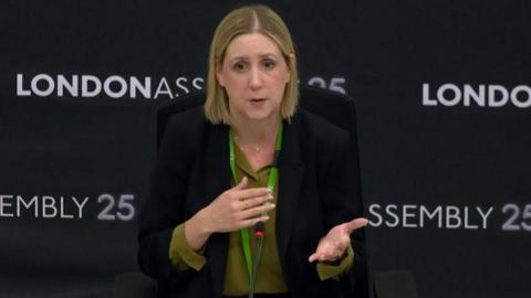 A blonde woman with a black jacket and green lanyard speaks at the London Assembly.