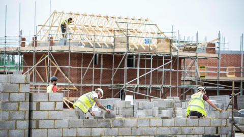 Several labourers lay a breeze block wall with an under-construction house with roof timbers and scaffolding behind them.