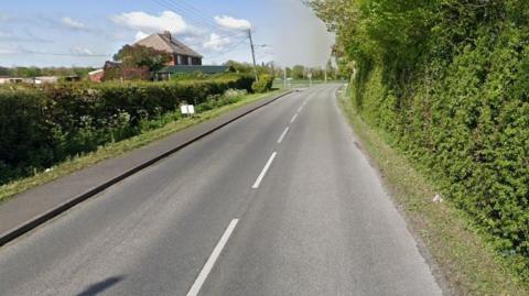 A road with a bush along the left-hand side and tree-lined down the right. In the distance on the left is a house.