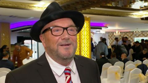 George Galloway wearing a black hat and a red, white and blue striped tie stands in front of a room full of white chairs with people talking to each other behind him.