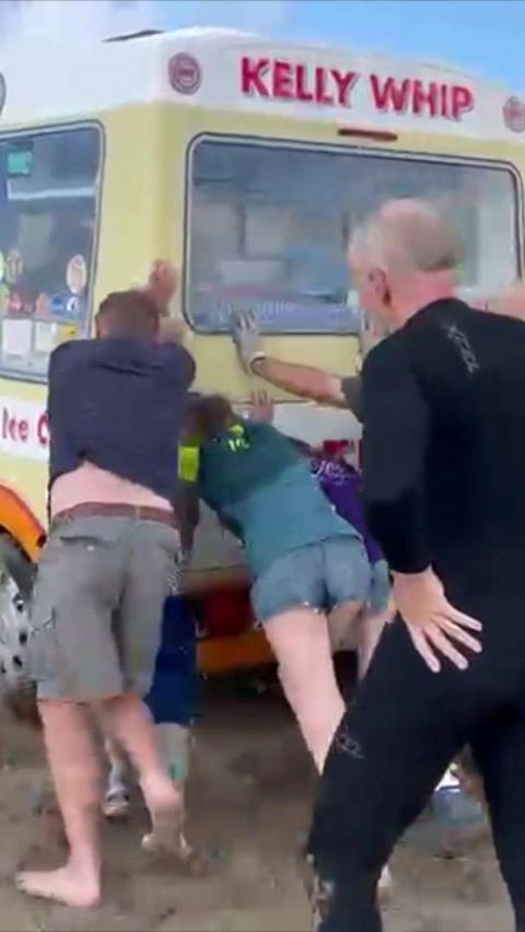 A group of people are pushing an ice cream van out of the sand.