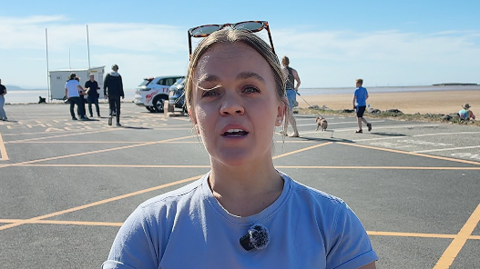 Ellie Simmonds with blond hair in a pony tail wearing a blue t-shirt and sunglasses resting on her head stands on the car park by West Kirby beach. 
