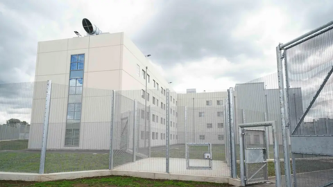 HMP Fosse Way is pictured behind a tall fence. It is a tall white-cladded building.