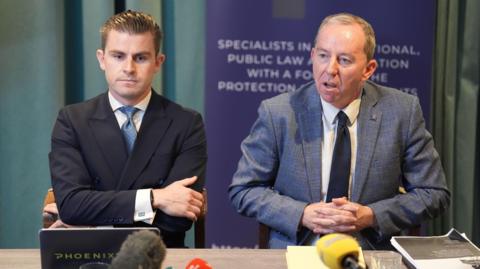Darragh Mackin and Peter Corrigan sitting behind a table. Darragh is on the left of the photo. He has short brown hair and is wearing a white shirt, blue tie and navy suit. He has his arms crossed. Peter is on the right of the photo and he is wearing a white shirt, navy tie and grey suit. 
