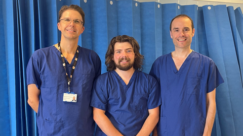 Prof Gavin Morley (centre) with PhD student Alex Newman (right) and surgeon Stuart Robertson. All three are wearing blue medical scrubs and standing in front of a blue hospital curtain