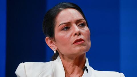 Priti Patel has black hair and looks pensive in her expression. She is wearing a white jacket.