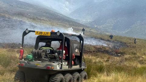 A small all-terrain vehicle being driven towards an area of smouldering moorland where firefighters are damping down