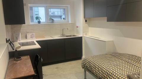 Photograph of one of the rooms at the Embassy homeless village in Castlefield. The image includes cupboards, a bed, a cooker and a kitchen sink.