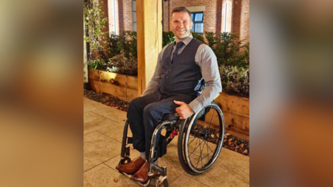 A man in his 40s in a wheelchair, wearing a suit and brown shoes. He is smiling and is outside by a planted area. 