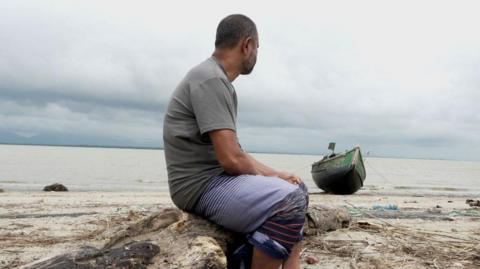 Nasir sits on a log on the bank of a river looking out across the river. His face is turned away and is not visible. He is wearing a grey t-shirt, shorts, and sandals. The day is overcast, and a small boat is also visible on the bank of the river. 