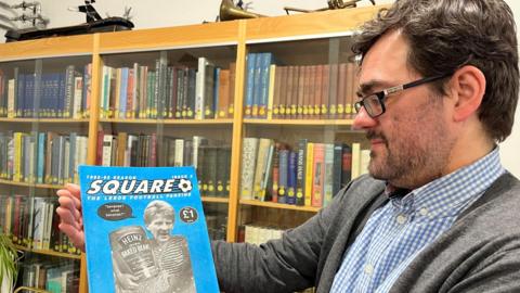 A picture of librarian Antony Ramm who is holding a fanzine with the words 'Square The Leeds United Football Fanzine'. Mr Ramm is wearing glasses and a blue and white checked shirt with a grey cardigan. In the background there are books on display in a cabinet. 