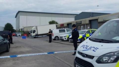 Image of a car park cornered off by police. Two police cars and a police van are parked along the side of the cark park, and two officers are standing in front of them. 