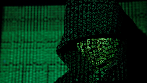 A hooded figure in darkness with Matrix-style green code projected on to their face and a wall behind them