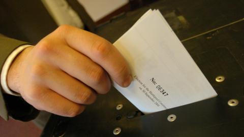 A hand, with a brown jacket visible on the sleeve, holds a folded white ballot paper in his thumb and forefinger.  It is just above the slot on a black ballot box. 
