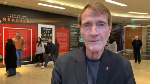 A man in a black suit jacket with a small red poppy badge on it, looks directly at the camera. Behind him are large displays, including one that says How do you measure up to Reacher?