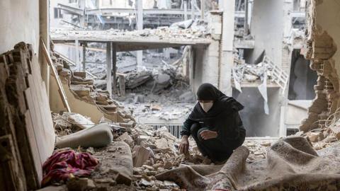 A woman wearing a black headscarf, dark hoodie and a white face mask crouches among thick concrete rubble and dust inside a heavily damaged building. The walls around her are blown open, exposing twisted metal and collapsed floors across multiple storeys. A patterned rug and scattered personal belongings are partially buried in the debris.