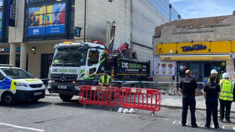 A lorry is seen from the height of the road after having crashed into a wall. Police officers are standing nearby.