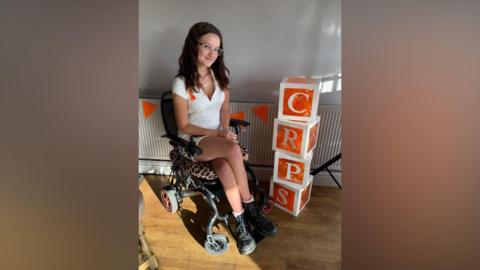Maddie, wearing a white dress, is sitting in a wheelchair indoors.