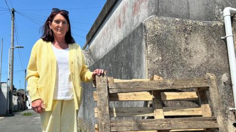 A woman is standing on a back street beside a wooden pallet that is leaning against a concrete wall. She is dressed in a yellow cardigan with a white top and yellow trousers. 