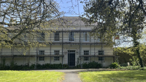 St Saviours Rectory. It's a large 2 storey building with a straigh path leading up to the door, with green grass and trees either side. There is scaffolding of the whole front of the building.