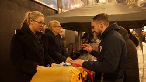 Three individuals engage in a discussion at an outdoor stall, surrounded by food and clothing items.