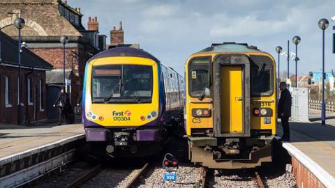 Two trains are stopped side by side at Grimsby station platform. The train on the left has a purple and yellow livery with the number 170304 visible on the front, while the train on the right is yellow and black with the number 153316. Both trains are positioned on parallel tracks, with station buildings on the left and a platform with modern lamp posts on the right. A man is standing on the right platform waiting for the train doors to open.