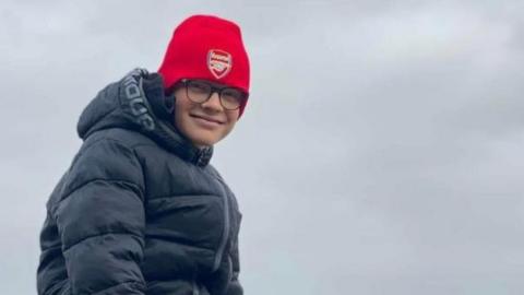 Harry Basham is sitting on a rock on a beach. He is wearing a red hat and a black hooded jacket. He is wearing black tracksuit bottoms and black and red trainers. The picture was taken on a cloudy day.