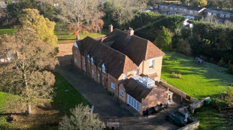 A large detached house surrounded by gardens in Marston Moretaine. There is a church tower and houses in the background. It is an aerial view picture.