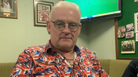 A man in a Union Jack shirt relaxes in a pub environment and smiles for the camera