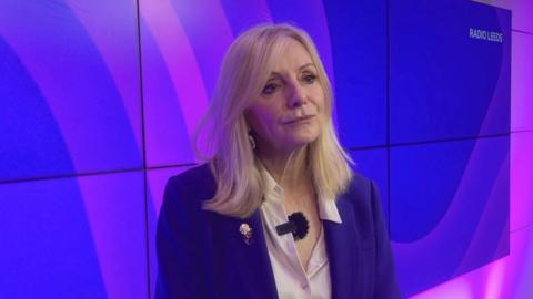 A woman wearing a purple blazer with mid length blonde hair stands in front of several tv screens