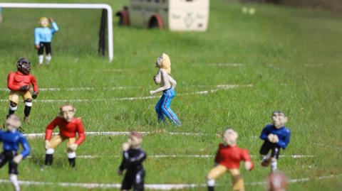 A close up of a football pitch with miniature model players. A figure of a topless blonde-haired woman can be seen running across the pitch.